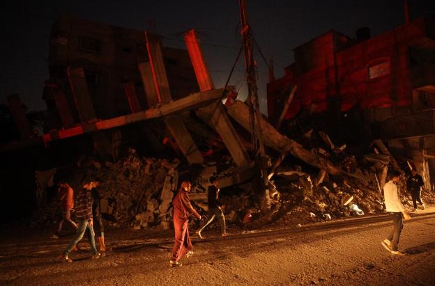 Palestinos pasan junto a un edificio destruido en el campo de refugiados de Al-Bureij, en el centro de la Franja de Gaza, durante la noche.