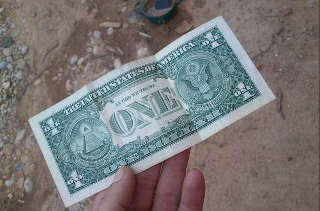 billete de un dólar sostenido por una mano, fondo piso de tierra 