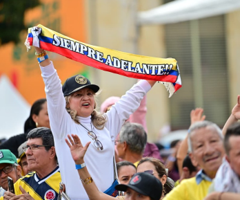En un evento masivo en plaza pública, una señora levanta una cinta con la bandera de Colombia que dice "siempre adelante"