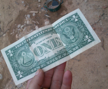 billete de un dólar sostenido por una mano, fondo piso de tierra 