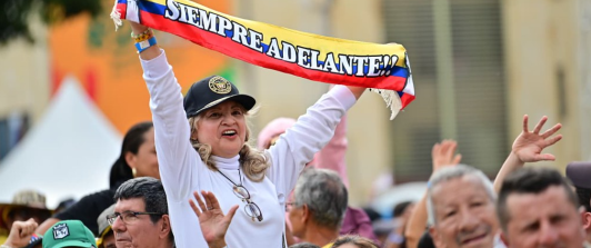 En un evento masivo en plaza pública, una señora levanta una cinta con la bandera de Colombia que dice "siempre adelante"