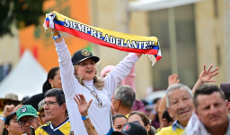 En un evento masivo en plaza pública, una señora levanta una cinta con la bandera de Colombia que dice "siempre adelante"