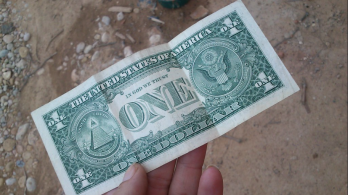 billete de un dólar sostenido por una mano, fondo piso de tierra 