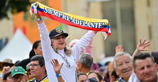 En un evento masivo en plaza pública, una señora levanta una cinta con la bandera de Colombia que dice "siempre adelante"