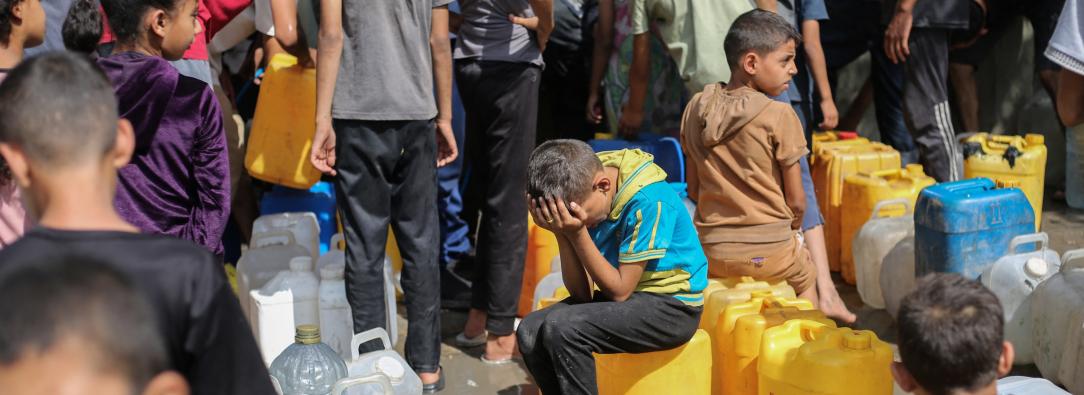 Niños palestinos sentados sobre sus contenedores mientras hacen cola para recibir agua en un punto de distribución benéfico en el campo de refugiados de Nuseirat, en la Franja de Gaza, sitiada por Israel.