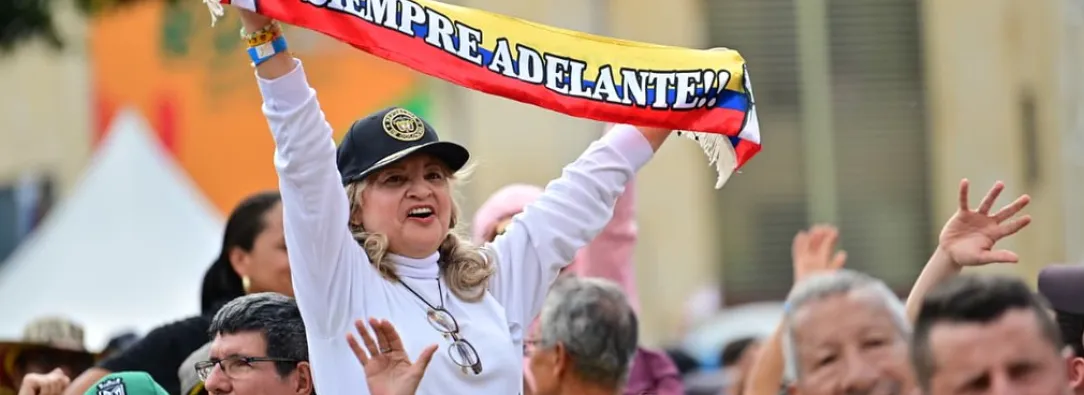 En un evento masivo en plaza pública, una señora levanta una cinta con la bandera de Colombia que dice "siempre adelante"