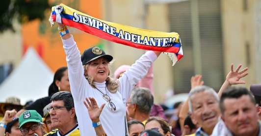 En un evento masivo en plaza pública, una señora levanta una cinta con la bandera de Colombia que dice "siempre adelante"