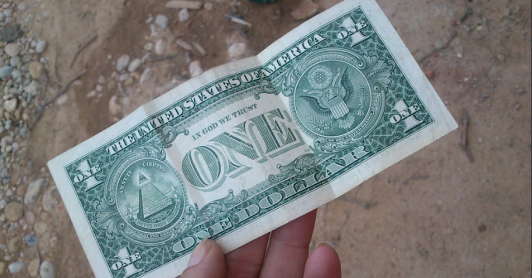 billete de un dólar sostenido por una mano, fondo piso de tierra 